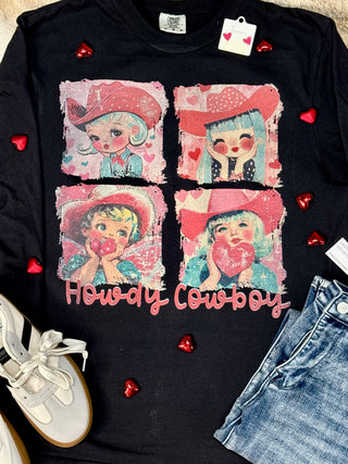 The Howdy Cowboy Graphic Tee by Western Edge Boutique features a vintage design of four cartoon cowgirls in pink cowboy hats holding hearts. "Howdy Cowboy" adds a Valentine's mood, with red heart beads, white sneakers, and blue jeans completing the look.