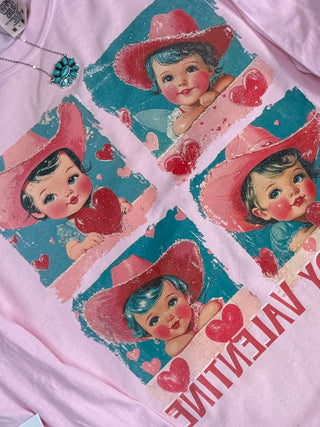 The Howdy Valentine Graphic Tee by Western Edge Boutique features a vintage-style design with four images of a baby in a pink cowboy hat, each holding a red heart, and "MY VALENTINE" at the bottom. Paired with a turquoise floral necklace, it’s ideal for Valentine's Day.