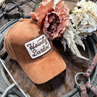 The "Howdy Darlin" brown corduroy hat from Western Edge Boutique is elegantly embroidered with the phrase "Howdy Darlin'" and adorned with pink and white flowers. It is displayed on a coiled rope, complemented by leather straps, creating a charming rustic western theme.