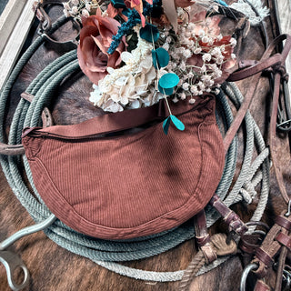 A brown, crescent-shaped Corduroy Cross Body from Western Edge Boutique mingles with coiled ropes and decorative floral arrangements. Nearby, a Howdy Darlin hat and metal horse bit sit on a textured surface, evoking a sense of rustic charm.