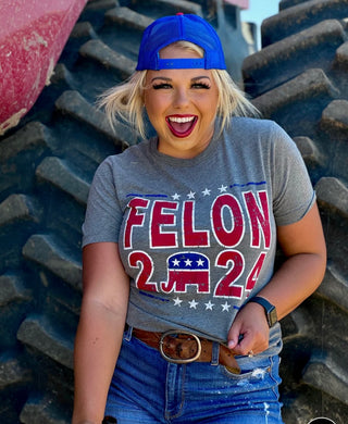 A smiling individual wearing the "Felon 2024 Tee" from Western Edge Boutique, along with a blue backward cap and gray T-shirt that reads "FELON 2024" with a Republican elephant emblem, stands in front of large tractor tires, hands on parts of their shorts and belt. The edgy style is reminiscent of high-demand Trump shirts found in aftermarket collections.