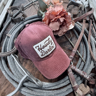 A dusty rose corduroy hat from Western Edge Boutique, featuring the embroidered words "Howdy Darlin'," sits among coiled ropes, leather straps, and a peach-colored artificial flower to create a rustic western ambiance. This delightful accessory perfectly complements any country-inspired outfit.