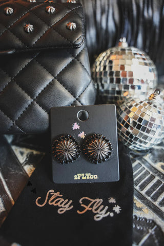 A close-up shot of a pair of "Giddy Up Concho Stud Midnight" earrings from Western Edge Boutique on a card labeled "2FLYCo". The striking black sunburst design earrings are displayed against a quilted black purse. In the background, there are two small disco balls and a black pouch with "Stay Fly" written in white.