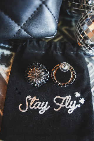 A close-up photo showing two earrings placed on a black drawstring bag. One earring is the "Giddy Up Concho Stud Midnight" from Western Edge Boutique, featuring a round, studded design, while the other has a round hoop with small black squares. The bag has the words "Stay Fly" embroidered in white, adding an edgy flair to these distinct jewelzzz.