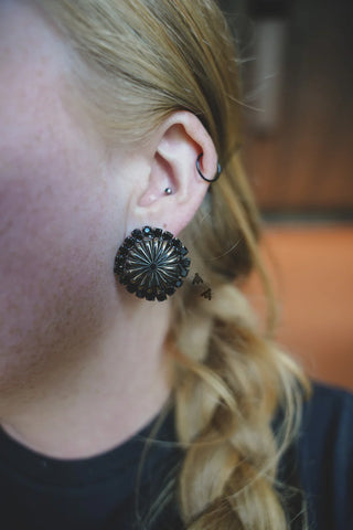 Close-up of a person's ear adorned with the Western Edge Boutique's Giddy Up Concho Stud Midnight, a large, ornate earring shaped like a flower with an edgy flair. Above it, a small stud piercing is paired with a black hoop earring higher on the ear. Light blonde braided hair cascades down, complementing the black garment they wear.