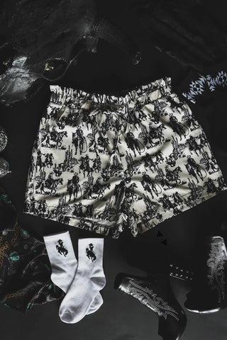 A pair of GONE WITH THE WINDS black and white patterned paperbag shorts with a jungle design from Western Edge Boutique laid on a dark surface. Surrounding items include white logo socks, shiny black shoes, and textured dark clothing.