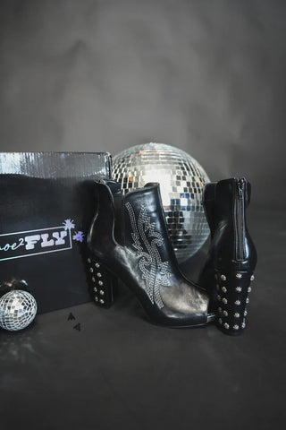 A pair of HIGH DEMAND *LUX black heeled ankle boots from Western Edge Boutique, featuring intricate embroidered designs on the sides, is displayed in front of a shiny black box labeled "Fly." Crafted from premium black leather, these boots boast studded heels. Two disco balls of different sizes are placed beside and behind them.