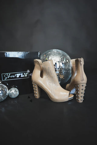 A pair of River Rock Western Heels by 2 Fly, featuring studded heels, a side zipper, and decorative stitching in tan leather, are displayed in front of a Shoefly box amidst large and small disco balls against a dark background.