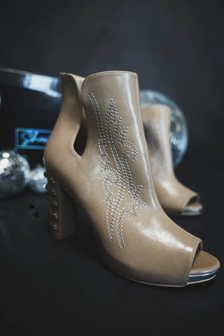 The River Rock Western Heels by 2 Fly are showcased against a dark backdrop, revealing their stylish tan leather peep toe design with intricate white stitching. The high-heeled boots feature stud detailing at the back. The scene is accented by reflective decorative spheres and a partially visible shoebox in the background.