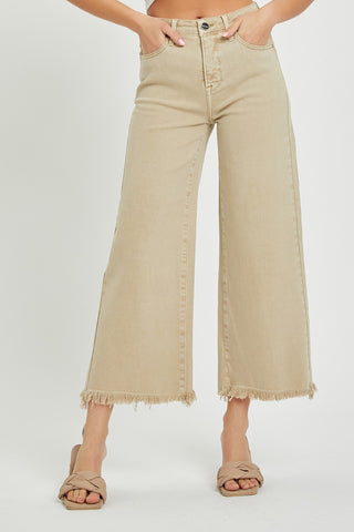 A person wears Western Edge Boutique's Sandy High Rise Crop Wide Leg Pants, which feature frayed hems and a beige hue. The high-waisted jeans provide excellent tummy control and come with front pockets and a button fly. Complementing the outfit, the person has on tan sandals with crisscross straps against a plain white background.