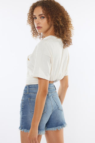 A woman with curly hair is wearing a light-colored t-shirt and chic Haven High Rise Distressed Mid Shorts from Western Edge Boutique. She is standing sideways, looking over her shoulder towards the camera against a plain white background.