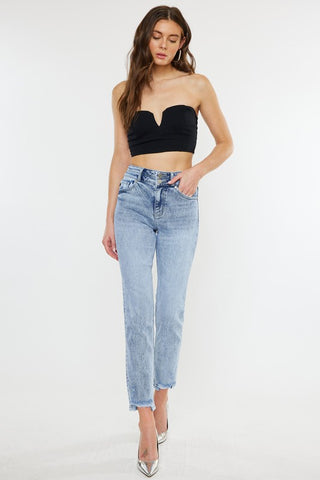 A woman with long, wavy brown hair is modeling the Kan Can Leona High Rise Slim Straight Jeans in a light stone wash. She pairs them with a black strapless crop top and metallic silver pointed-toe heels. Posing against a plain white background, she has one hand casually placed in her pocket.