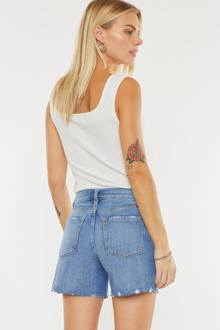 A woman with long blonde hair is standing and looking to the side, wearing a white sleeveless ribbed top, Kan Can's Nellie High Rise Stone Wash Shorts in 100% cotton blue denim, and earrings. She has a floral tattoo on her upper right arm and is holding her left arm behind her.