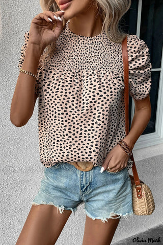 A person wears the Olivia Mark Khaki Leopard Print Blouse by Western Edge Boutique, featuring ruched puff sleeves, paired with light blue denim shorts. They carry a woven rattan bag and accessorize with a gold bracelet and ring, standing against a white wall.