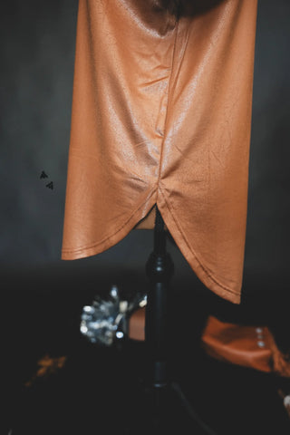 A close-up of the lower part of the Swanky Faux Leather Cotton Shirt by 2 Fly, made from a shiny, brown stretchy leather lux material, displayed on a black mannequin stand. The fabric features a soft sheen and a pointed asymmetrical hem. Blurred objects in the background suggest a studio setting.