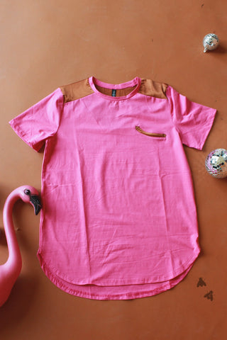 The Dolly Approved Satin Shoulder Tee by 2FLY, a pink short-sleeved Western top with brown satin shoulder accents, is laid on a flat brown surface. A small zippered pocket detail is visible on the chest. To the left of the shirt, there's a pink flamingo decoration, and two small disco ball ornaments add a retro rodeo vibe on the right.