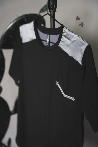 The Dolly Approved Satin Shoulder Tee by 2FLY, in black with light gray satin patches on the shoulders, hangs on a hanger. Crafted from solid fabric, the shirt features a small left-side pocket embellished with a design resembling two arrows pointing downward. In the background, two flies are visible but slightly out of focus.