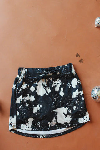 The Galuxy Hide Skort by 2 Fly, featuring a black and white tie-dye pattern, is crafted from stretchy fabric and equipped with an elastic waistband and drawstring. It lies flat against a brown surface, while two small disco balls in the bottom right corner of the image add a touch of sparkle.