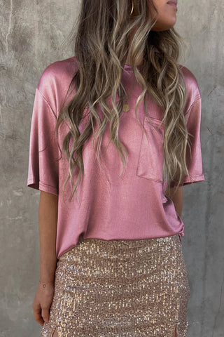 A woman with long, wavy hair is wearing the loose, shiny pink Luca Satin Short Sleeve Pocket Blouse from Western Edge Boutique and a beige sequin skirt. She is posed against a gray textured background and is looking to the side, with her right arm resting by her side, showcasing her chic boyfriend fit ensemble.