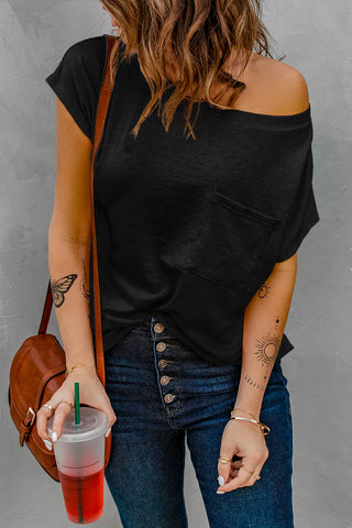 A woman with wavy hair sporting the Bellarose Pocket Tee with Side Slits from Western Edge Boutique and blue jeans holds a red iced drink. Her brown bag complements butterfly and sun tattoos, while bracelets adorn her wrists against a textured gray wall.