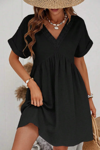 The woman is wearing the Arrow Black V Neck Dress from Western Edge Boutique, styled with a straw hat. Her look is accessorized with a shell necklace and bracelets, and she has wavy hair. The cotton dress features a casual, loose fit and lace details on the neckline. She stands in a light-colored room.