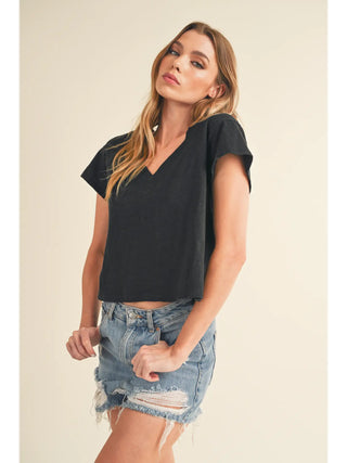 A woman with long hair poses against a plain backdrop, wearing the Rowen V Neck Black Women's Casual Top by Western Edge Boutique and frayed denim shorts. The 100% cotton outfit complements her relaxed stance as she holds her shorts at the sides, looking at the camera neutrally.