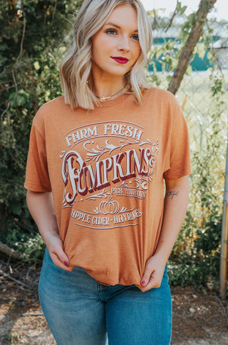 A person with blonde hair and red lipstick is standing outdoors in front of green foliage. They are wearing a true-to-size brown Fall Tee from One24rags, made from a comfortable cotton-poly blend, featuring white text that reads, "Farm Fresh Pumpkins - Pick Your Own - Apple Cider - Hayrides," paired with light blue jeans.