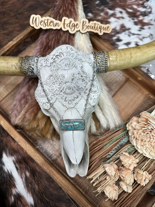 A decorative display featuring a cow skull adorned with intricate lace and the Marjorie Marbled Turquoise Bar Necklace by Wild Junkie. It is framed by rustic wooden elements and artificial flowers, with a sign reading "Western Edge Boutique" placed above the skull, set against a background with cowhide patterns.