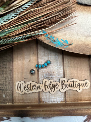 A rustic wooden surface showcases the Harriet Turquoise Ear Climber Earring Set, featuring silver ear climbers adorned with turquoise gemstones and a small sunburst charm. Nearby are natural feathers with brown and turquoise accents, alongside a wooden sign that reads "Western Edge Boutique" in decorative script, capturing an authentic western flair.