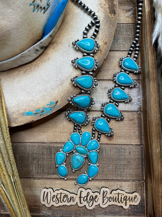 The Monroe Turquoise Squash Blossom Necklace, featuring rustic teardrop and oval turquoise stones set in an antique silver plate frame, lies on a wooden surface, partially covering a beige cowboy hat with blue accents. The brand name "Western Edge Boutique" is inscribed at the bottom on a wooden plaque.