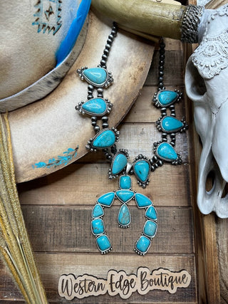 A rustic display featuring the Maribel Turquoise Squash Blossom Necklace with intricate silver detailing and a stunning squash blossom pendant laid on a wooden surface. Nearby are a beige cowboy hat with turquoise accents, a cow skull, some wheat stalks, and a wooden sign reading "Western Edge Boutique.