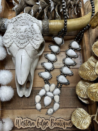 A decorative skull adorned with lace is placed on a wooden surface surrounded by rustic decorations. Beside it is an antique silver plate holding the Hope White Squash Blossom Necklace from Western Edge Boutique, featuring large, white stones. At the bottom, a sign reads "Western Edge Boutique.