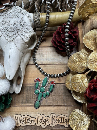 A rustic, Western-themed display showcases the Ripley Navajo Style Cactus Pendant Necklace, adorned with silver Navajo-style beads and a cactus-shaped pendant featuring green stones and small red accents. The setup includes a decorative skull, pinecones, braided rope, and golden ornaments on a wooden background. The text "Western Edge Boutique" is visible at the bottom.