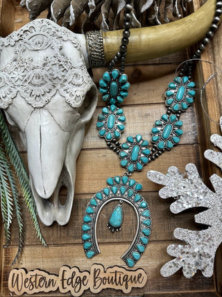 A decorative display showcases the Mavery Turquoise Squash Blossom Necklace by Western Edge Boutique, featuring multiple oval turquoise stones set in silver arranged around a silver horseshoe shape. The setup includes an Antique Silver Plate, a cow skull, a lace skull cap, a large horn, and silver glittery ornaments. Text reads "Western Edge Boutique.