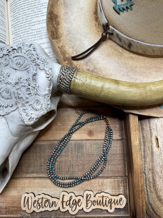 A decorative western-themed display featuring a partially open book, a cow skull adorned with lace, a hat with a burnished patina and decorative band, the Nadine Navajo Style Patina Layered Choker by Western Edge Boutique, and a Western Edge Boutique sign on a wooden surface.