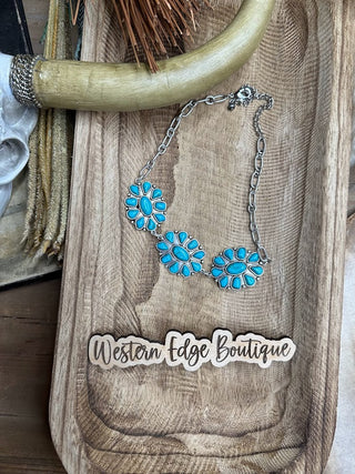 A Jo Turquoise Pendant Necklace with flower-shaped turquoise charms in antique silver plate is displayed on a wooden tray. Nearby is a sticker with the text "Western Edge Boutique." The scene also includes a decorative horn and a fringed item in the background, all nickel free.