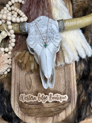 The Jayde Cactus Druzy Stone Necklace from Western Edge Boutique features a decorative white skull with horns, adorned with a lace headdress and mounted on a faux fur background. Wooden beads are draped over the horns, and the ensemble includes a versatile 16" necklace with a 3" extender. The "Western Edge Boutique" brand sign is placed beneath the skull.