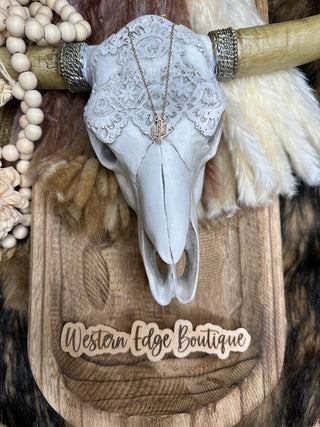 A decorative skull adorned with a lace headpiece and jewelry, featuring the Sharlene Hematite Druzy Cactus Necklace, hangs on a wooden plaque. Beaded garlands drape over the skull's horns. The plaque is engraved with "Western Edge Boutique." Fur and feathers surround the display, adding texture and rustic charm.