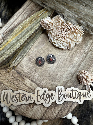 A pair of Odella Western Concho Stud Earrings featuring red centers are displayed on a wooden surface. Dried flowers and a wooden sign with cursive "Western Edge Boutique" lettering add a rustic touch to the arrangement.