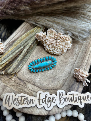 A turquoise brooch with small turquoise stones arranged in a symmetrical pattern is placed on a wooden tray. The tray, adorned with dried flowers and wheat stems, showcases a sign that reads "Western Edge Boutique" alongside the Marley Natural Stone Adjustable Ring.