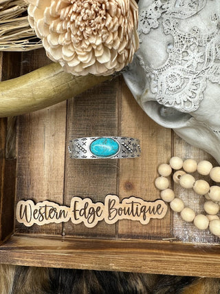 The Pacey Turquoise Cuff Bracelet from Western Edge Boutique is displayed on a wooden surface, surrounded by rustic decor including a faux flower, lace cloth, wooden beads, and a "Western Edge Boutique" sign. This natural stone bracelet features a striking turquoise gem set in antique silver plate.