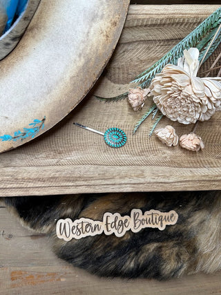 A rustic wooden display showcasing a weathered cowboy hat with a turquoise accent, dried white flowers, the Kendall Western Concho Hairpin featuring burnished silver with turquoise color highlights, and a wooden "Western Edge Boutique" sign sits atop a fur throw.