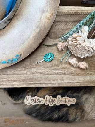 A rustic setting featuring a wide-brimmed hat with turquoise accents on the left, a turquoise gemstone brooch, dried flowers, and a branded "Western Edge Boutique" wooden tag. Amongst the items rests the delicate Lyndy Western Conch Hairpin by Western Edge Boutique. All items are beautifully arranged on a wooden surface.