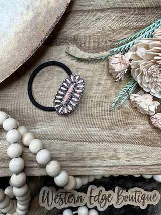 A wooden tray holds the Pixlynn Western Concho Hair Tie, featuring a decorative pink and burnished silver oval piece. Surrounding the hair tie are cream-colored wooden beads, dried beige flowers, and a sprig of greenery. Text at the bottom reads "Western Edge Boutique.