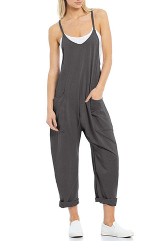 A woman is standing and wearing a loose-fitting, dark gray Charmo Randy Hotshot Onesie V-Neck Romper with rolled-up pant cuffs and a white tank top underneath. She has her hands in the romper's side pockets and is sporting white slip-on sneakers.