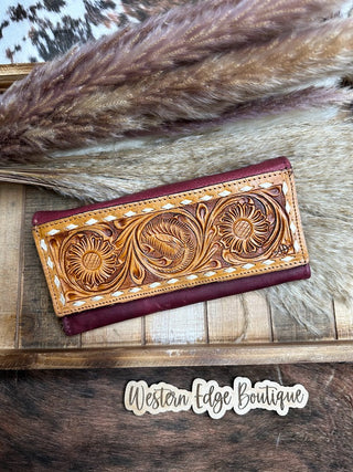 An intricately detailed, tooled wallet with floral designs in rich red leather from American Darling. This showstopper quality piece is placed on a rustic wooden surface with feathery dried plants in the background. The text "Western Edge Boutique" is visible at the bottom.