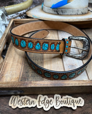 A unisex Parker Tooled Leather Belt by American Darling, featuring turquoise accents and an antique finish metal buckle, is displayed on a wooden tray. In the background, a cowboy hat adorned with a blue feather adds to the western fashion vibe. A wooden sign in front reads "Western Edge Boutique.