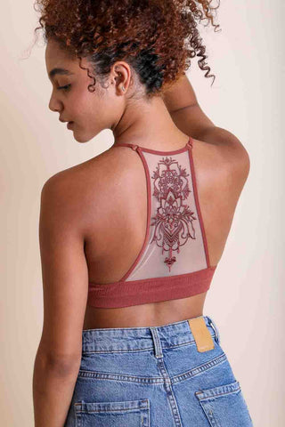 A person with curly hair is shown from the back wearing the Nicolette Tattoo Mesh Racerback Bralette by Leto Collection in a mauve color, featuring intricate lace detailing. The bralette has a sheer mesh back with an ornate design and no adjustable strap. The person is also wearing high-waisted blue jeans with a visible leather patch on the waistband.