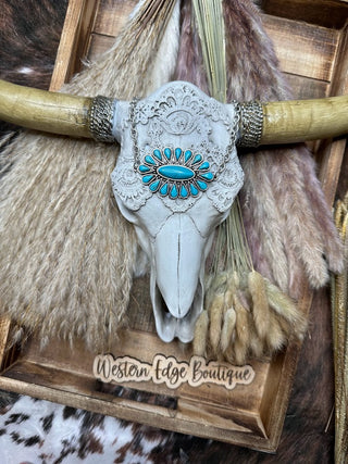 A decorative longhorn skull, adorned with the Farynn Turquoise Stone Necklace, is displayed on a wooden tray. The skull is surrounded by dried pampas grass and features engraved floral patterns. Below the skull, a sign reads "Western Edge Boutique.