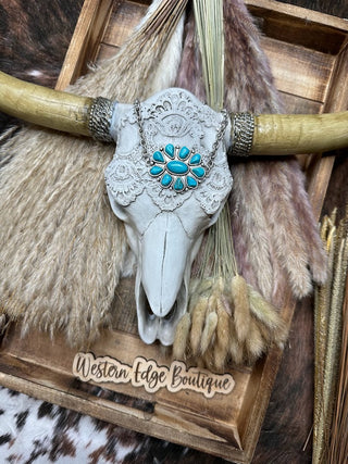 Against a rustic background, a decorative cow skull adorned with intricate lace is beautifully displayed. The skull is surrounded by pampas grass and other dried plants. An antique silver plate necklace, the Maelyn Turquoise Stone Choke Necklace, highlights the display beside a sign for Western Edge Boutique.